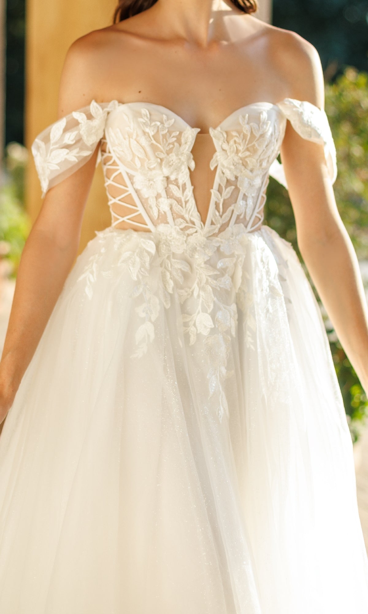 A woman wears the Long Formal Dress: Tina Holly TW480—an off-the-shoulder white bridal gown with lace detailing, floral appliques, a deep neckline, corset bodice, and flowing tulle A-line skirt.